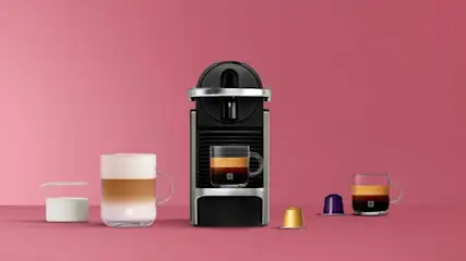 Zwarte Nespresso koffiemachine met twee glazen kopjes koffie en kleurrijke capsules op een roze achtergrond.