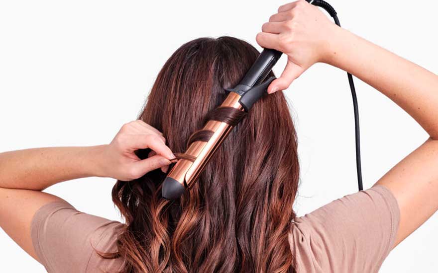 A person with brown hair is using a curling iron with copper-coloured plates to create curls against a white background.