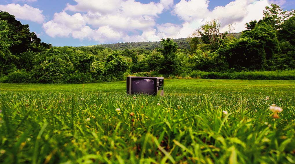 Een oude tv staat in een groen grasveld, met bomen en een blauwe lucht met wolken op de achtergrond.