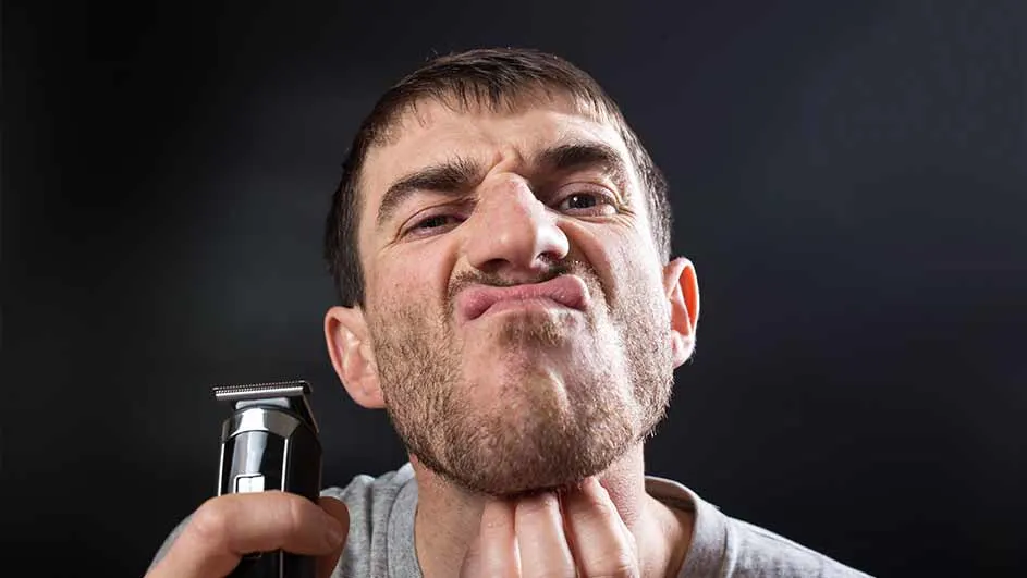 Een man trekt een gezicht terwijl hij een tondeuse vasthoudt en zijn kin aanraakt, tegen een donkere achtergrond.