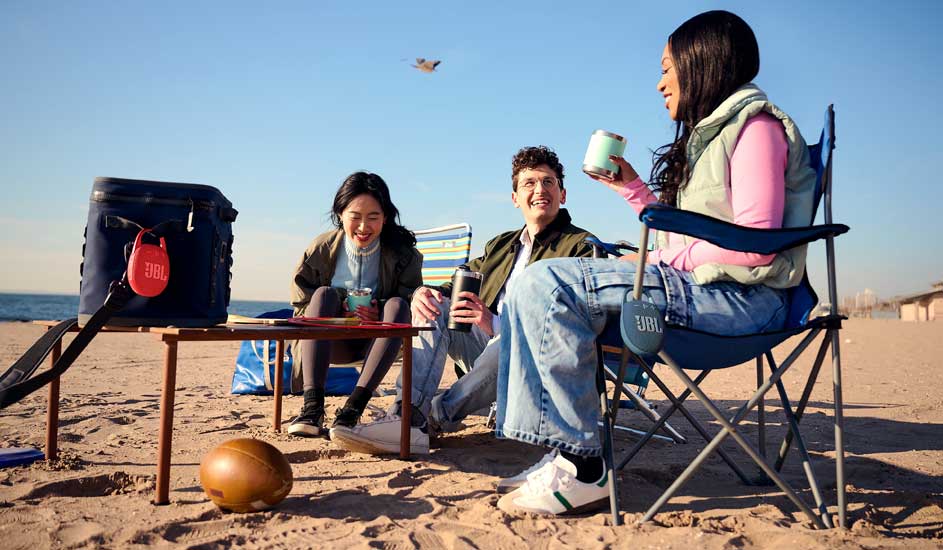 Drie vrienden zitten op het strand, lachen samen, drinken uit bekers naast een koelbox, terwijl de zee en een vogel op de achtergrond liggen.