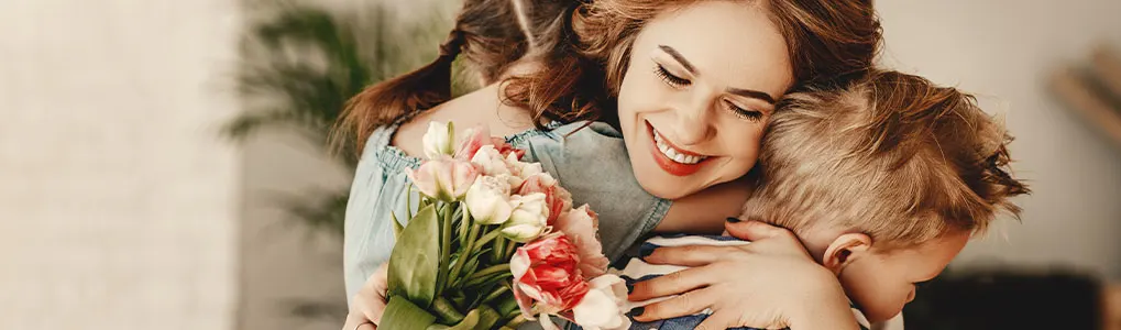 A woman in a blue shirt smiles, holding pink and white tulips, hugging two children in a light room.