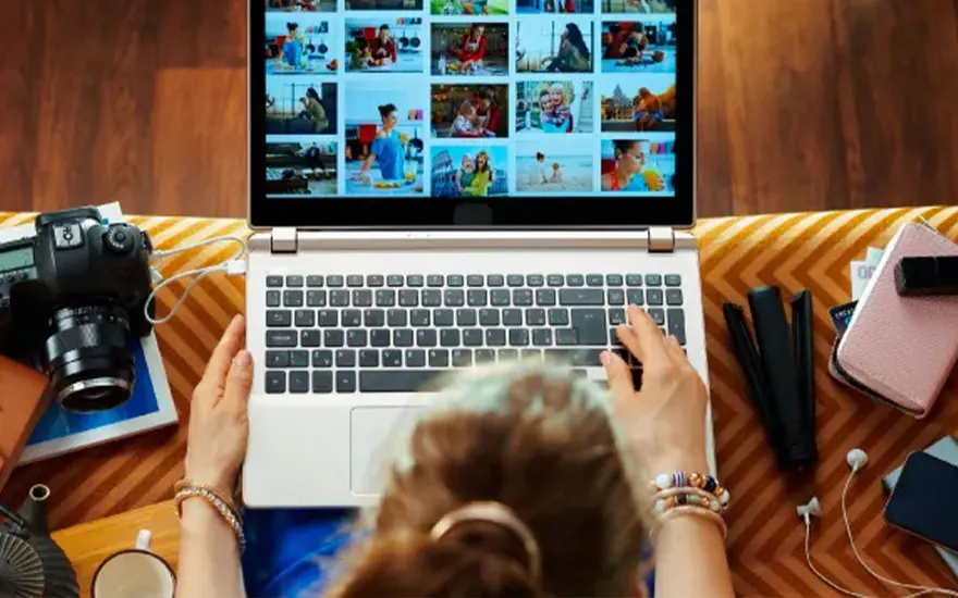 Vrouw typt op een laptop met foto's, naast een camera, notitieboek, telefoon en oordopjes op een gestreepte achtergrond.