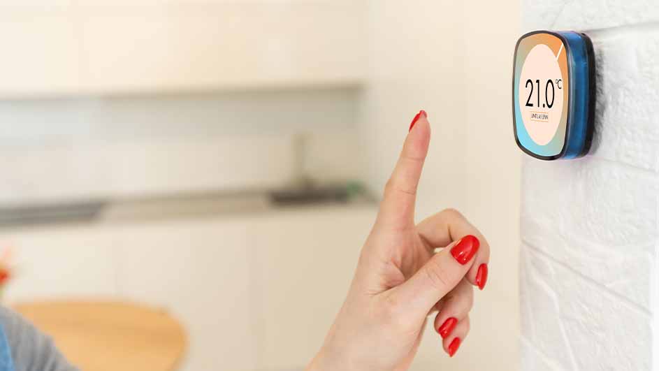 Een hand met rood gelakte nagels wijst naar een digitale thermometer aan een witte muur, met een keuken op de achtergrond.