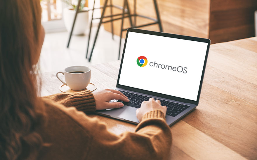 Vrouw typt op laptop met ChromeOS logo op scherm, draagt bruine trui, koffiekop naast laptop, café-achtige setting. 