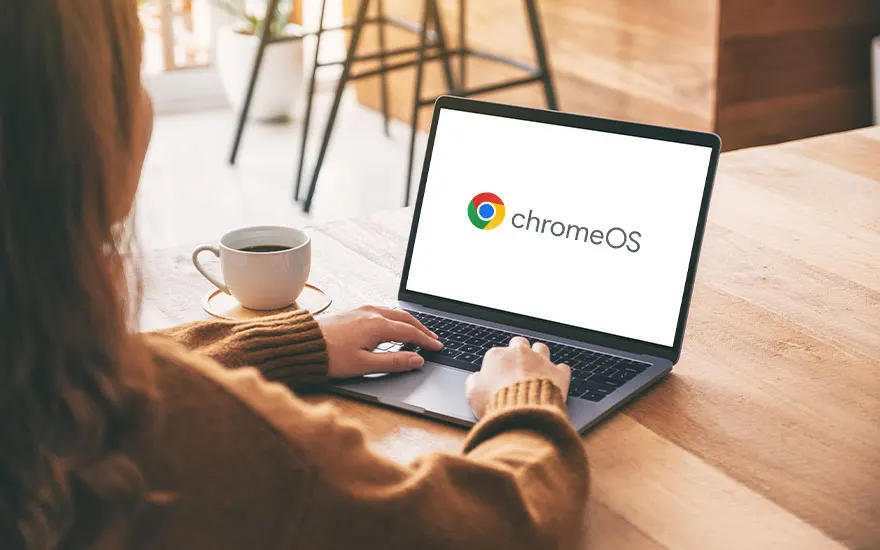 Vrouw typt op laptop met ChromeOS logo op scherm, draagt bruine trui, koffiekop naast laptop, café-achtige setting.