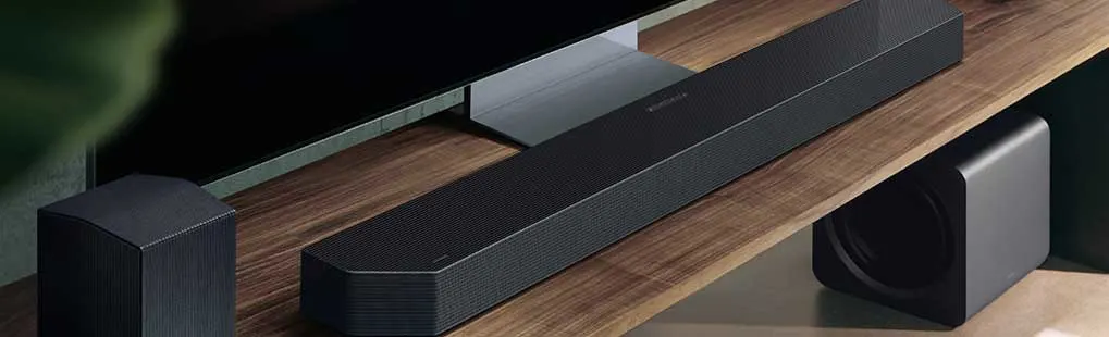 Op een houten tv-meubel staan een soundbar, twee achterspeakers en een subwoofer naast een grote flatscreen tv, allemaal in zwart design.