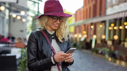 Een vrouw met een hoed gebruikt een telefoon in een stedelijke omgeving met gebouwen en verlichting op de achtergrond.