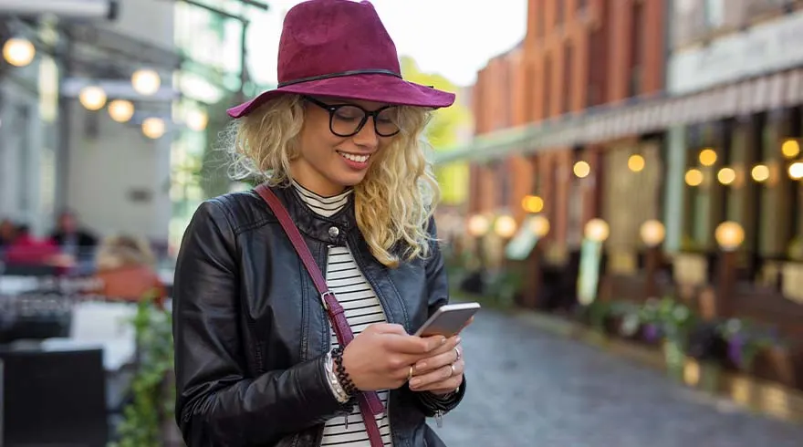 Een vrouw met een hoed gebruikt een telefoon in een stedelijke omgeving met gebouwen en verlichting op de achtergrond.