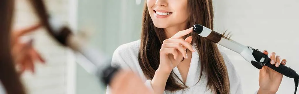 A smiling woman in a dressing gown curls her hair with a curling iron; the background is blurred, as if someone watches.