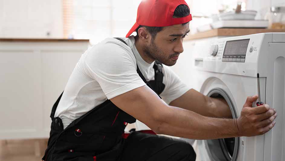 Man in a red cap and black overalls repairs a white washing machine with a screwdriver in a bright kitchen setting.