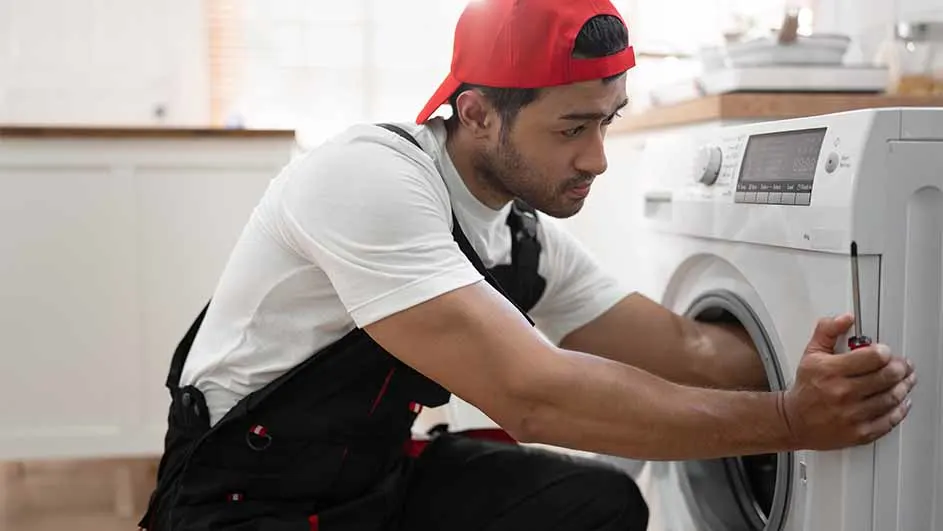 Man in a red cap and black overalls repairs a white washing machine with a screwdriver in a bright kitchen setting.