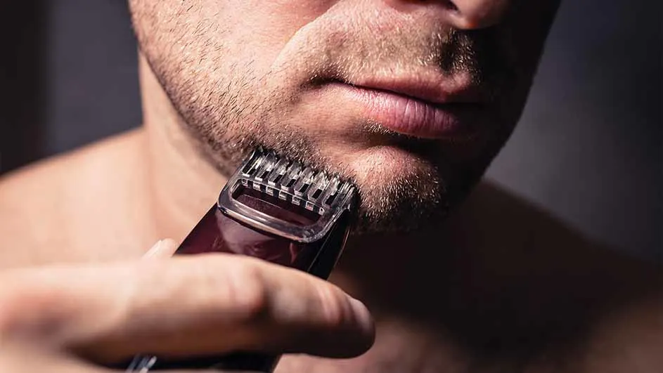 Een man trimt zijn baard met een donkerrode trimmer tegen een donkere achtergrond.
