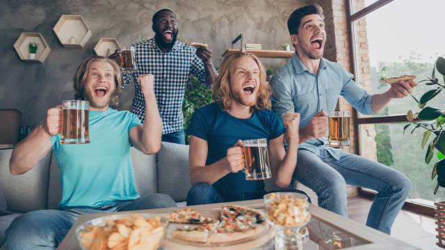 Vier mannen juichen met bier en pizza. Een tafel met snacks staat voor hen. Een raam en decoratieve planken zijn op de achtergrond.