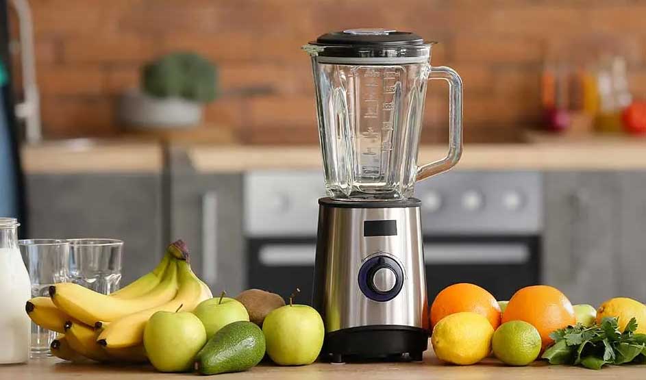 A blender sits on a kitchen worktop; in front are bananas, apples, citrus and herbs, with glasses and a bottle of milk beside it.