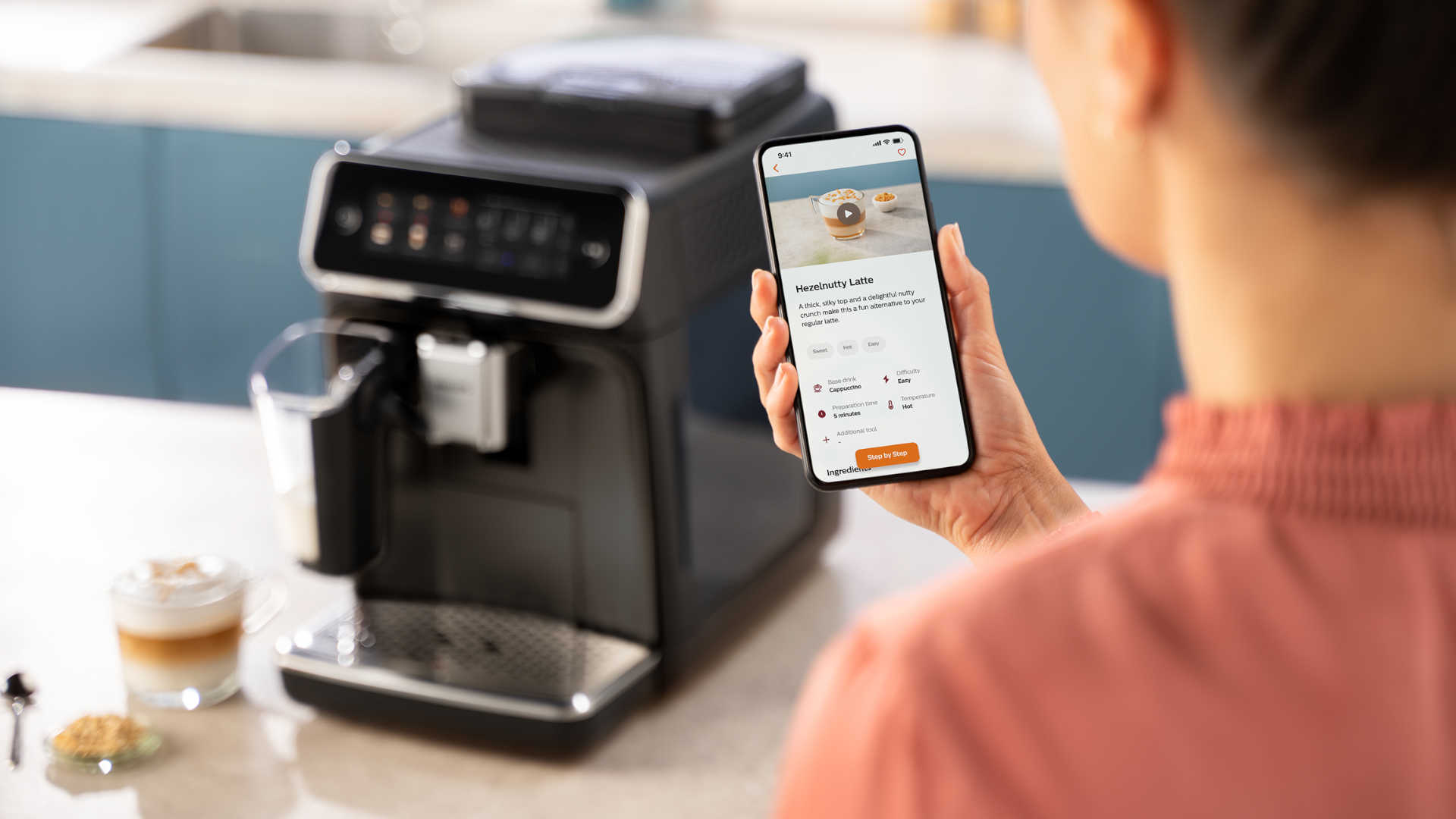 Vrouw bekijkt recept op een telefoon naast een koffiemachine. Er staat een glas latte en een koekje op de tafel.