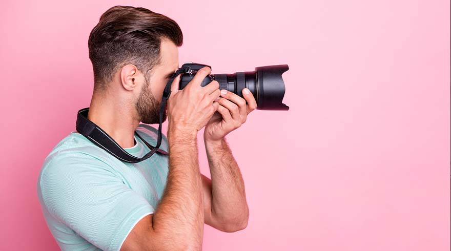 Een man met een camera tegen een roze achtergrond. Hij draagt een lichtblauw shirt en een zwarte camerariem.