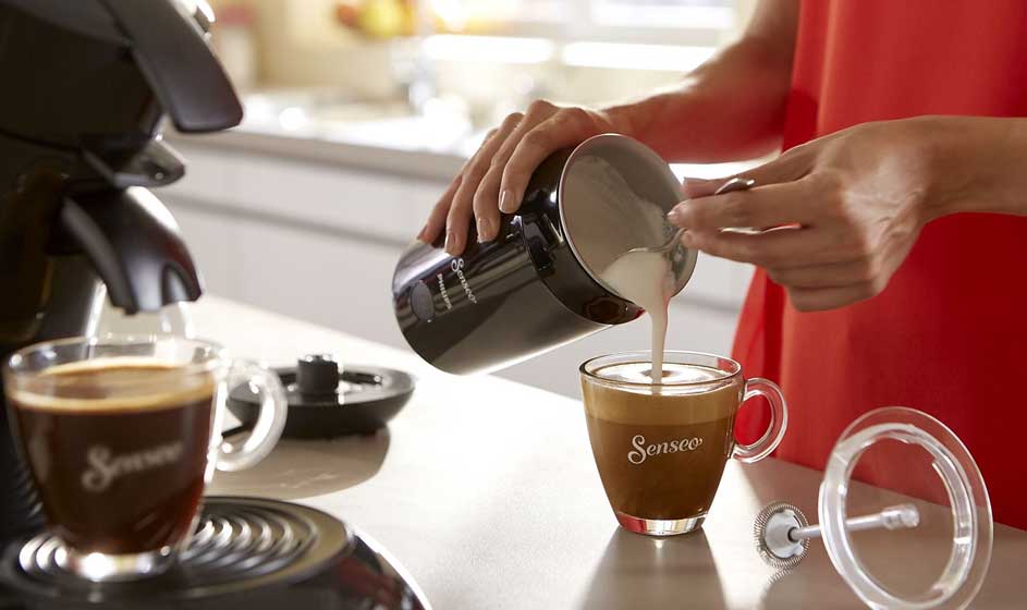 Een persoon schenkt warme melk uit een zwarte opschuimer in een Senseo-kop koffie, naast een machine en onderdelen op het aanrecht.