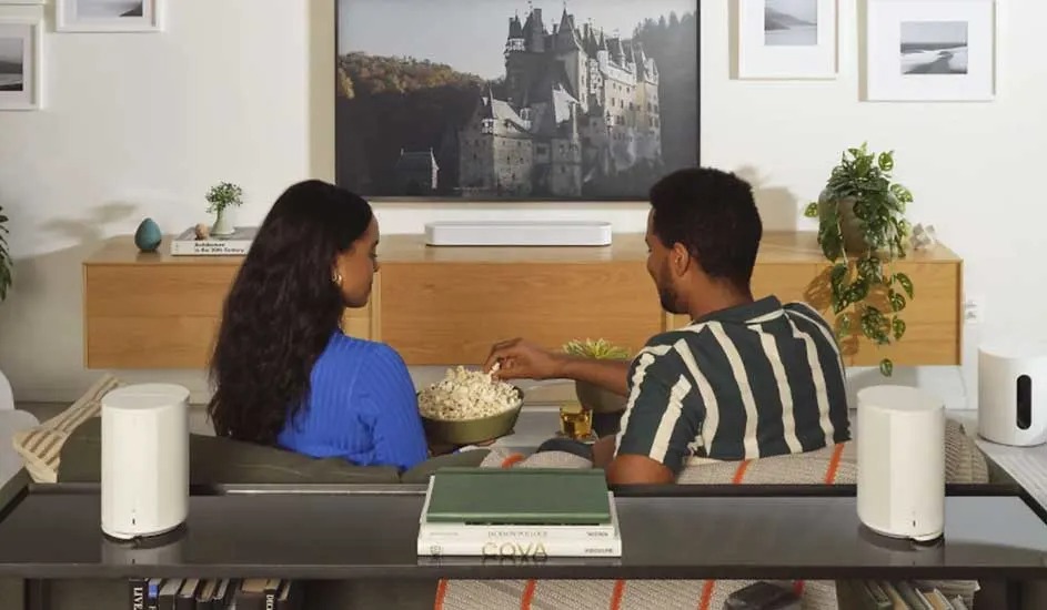 Twee mensen zitten op een bank en kijken tv met een kom popcorn. In de kamer staan meerdere witte Sonos speakers en planten.