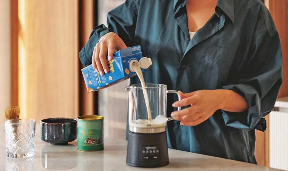 Een persoon giet melk uit een blauw pak in een kleine zwarte melkopschuimer, op een aanrecht met kopjes en potjes ernaast.