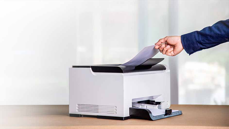 Een hand in een blauw overhemd legt een vel papier in een witte printer op een houten tafel.