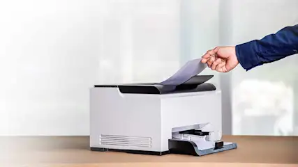 Een hand in een blauw overhemd legt een vel papier in een witte printer op een houten tafel.