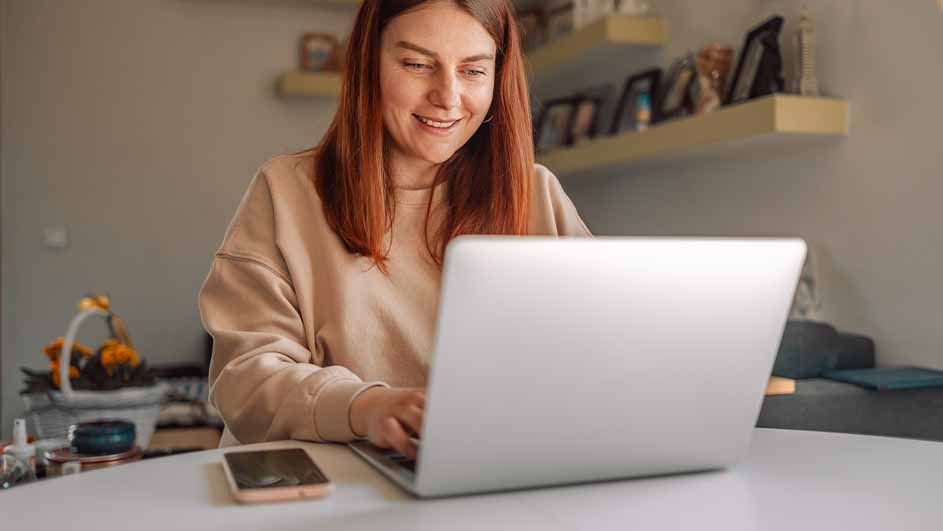 Een vrouw zit aan een witte tafel en typt op een laptop, met een smartphone ernaast. Op de achtergrond staan planken aan de muur.