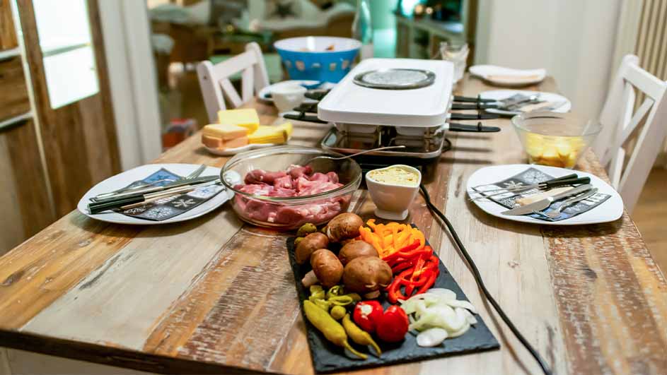 Houten tafel met raclette, borden, bestek, vlees, kaas, groenten en champignons. Stoelen zijn zichtbaar aan de randen.