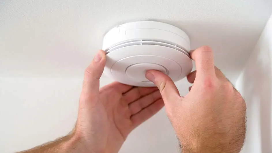 Twee handen bedienen een witte rookmelder aan een wit plafond, nabij een witte muur.