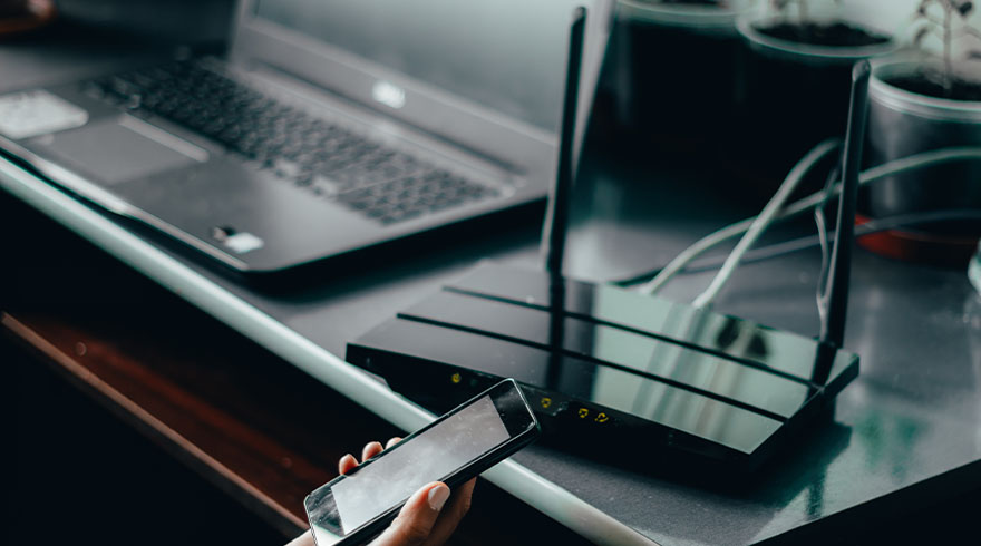 Een hand houdt een smartphone vast naast een laptop en een zwarte router met antennes op een tafel.