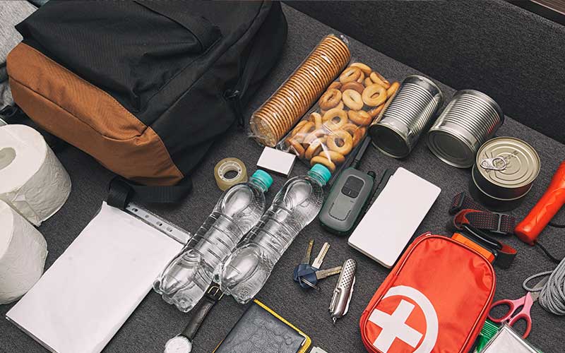 Laid-out emergency go-bag with water bottles, canned food, snacks, flashlight, first-aid kit, and assorted tools.