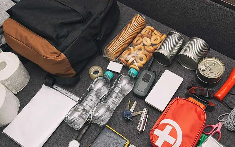 Laid-out emergency go-bag with water bottles, canned food, snacks, flashlight, first-aid kit, and assorted tools.