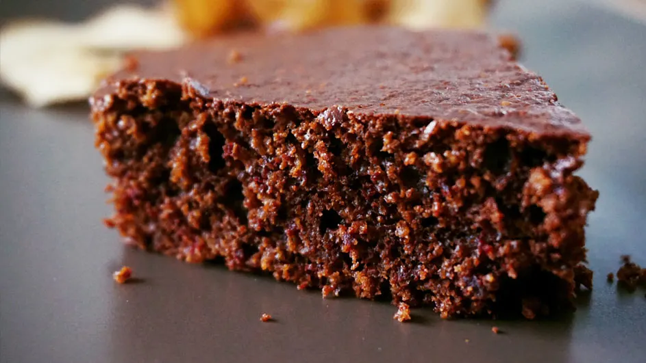 Een stuk chocoladetaart met glazuur op een grijs bord, tegen een vage achtergrond van gebakken stukjes.