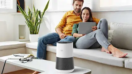 Een ontspannen koppel zit op een lichte sofa met tablet, terwijl op de salontafel een moderne luchtreiniger zachtjes de ruimte zuivert.