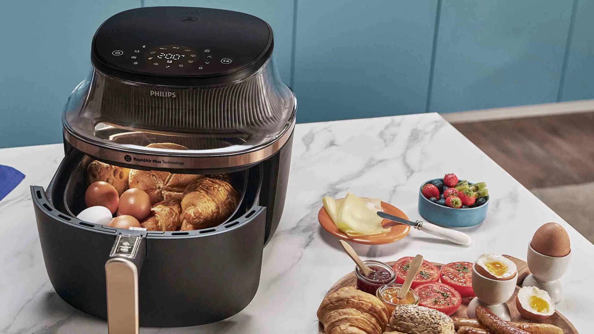 Een heteluchtfriteuse gevuld met eieren en croissants staat naast een bord met kaas, fruit en gekookte eieren op een marmeren tafel.