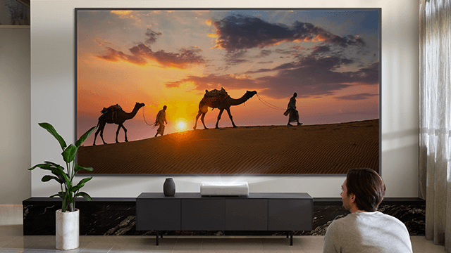 Een man kijkt naar een tv met een woestijnscène met kamelen en mensen tegen een oranje zonsondergang. Een plant en een tv-meubel staan in de kamer.