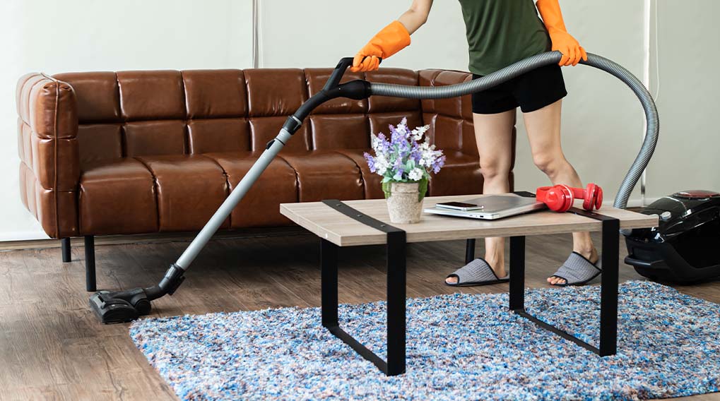 Een persoon stofzuigt een blauw vloerkleed, met een bruine bank en een tafel met bloemen en gewichten in de achtergrond.