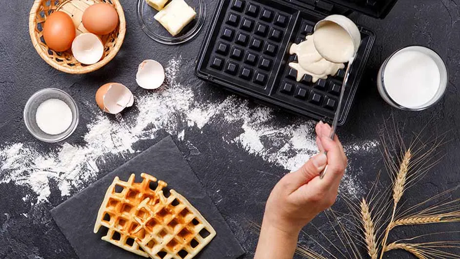 Wafels worden gemaakt. Ingrediënten zoals eieren, melk en bloem zijn aanwezig bij een open wafelijzer op een donkere ondergrond.