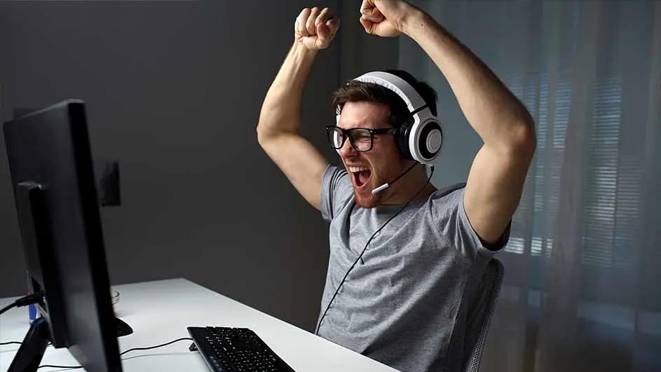 A gamer wearing a white headset and glasses throws his arms up in triumph, shouting at his monitor in a bright modern room.