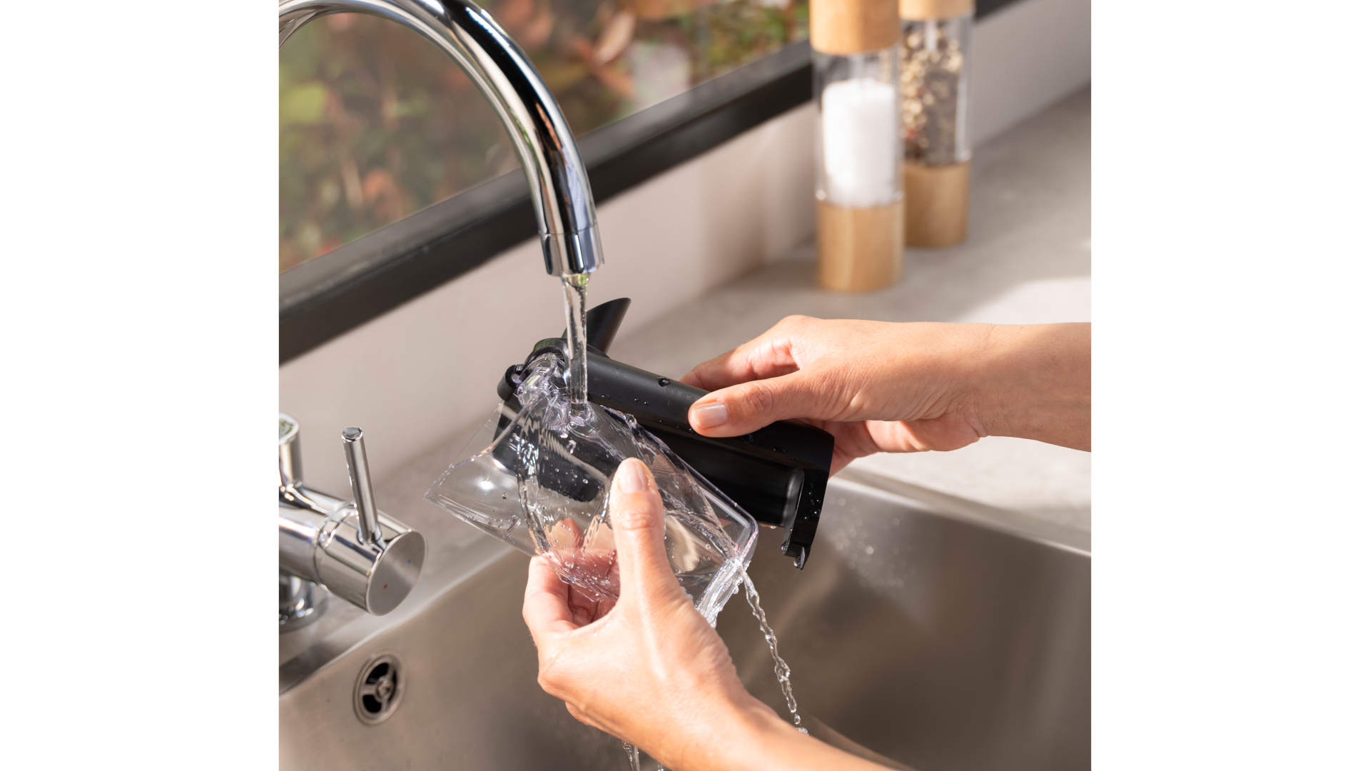 Iemand spoelt een doorzichtige beker en een zwart apparaat onder een zilveren kraan in een metalen gootsteen.