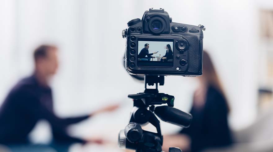 Een camera op een statief filmt twee mensen in een gesprek, met vage figuren op de achtergrond.