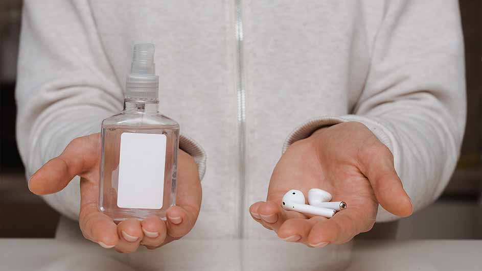 A person holds a clear hand sanitizer bottle in one hand and white wireless earbuds in the other. The background is neutral.