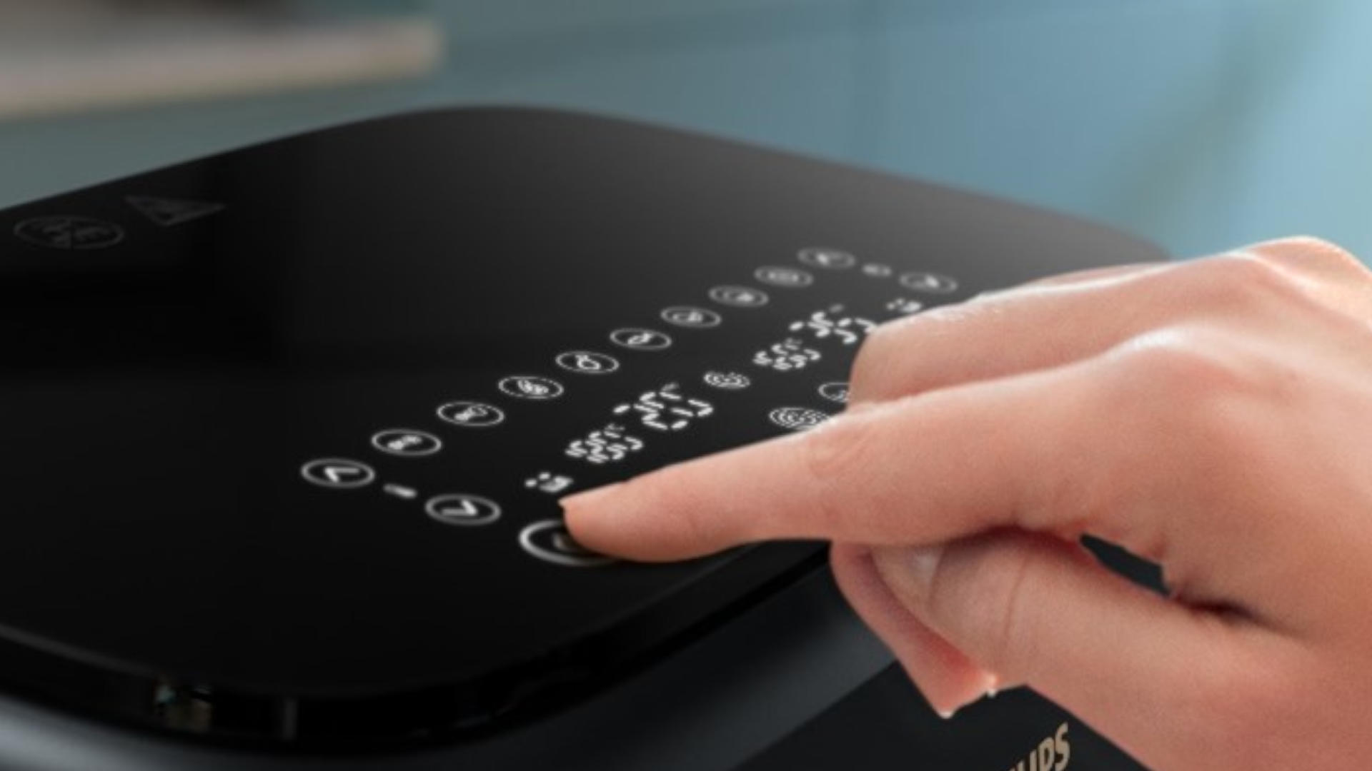 Een vinger drukt op de aan/uit knop van een zwarte Philips airfryer met een digitaal display tegen een blauwe achtergrond.