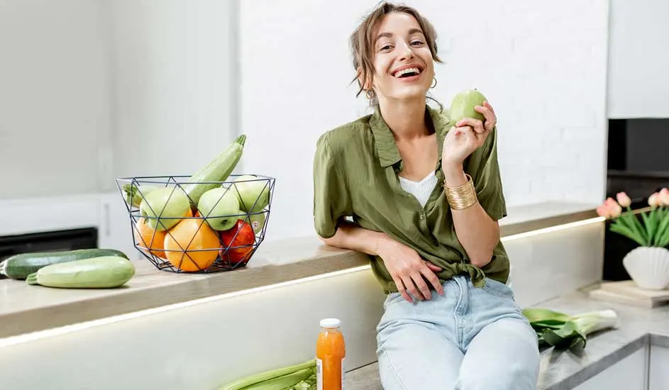 Op het aanrecht staat een metalen fruitmand met appels, sinaasappels, courgettes en paprika’s. Iemand eet een groene appel en draagt een groene blouse.