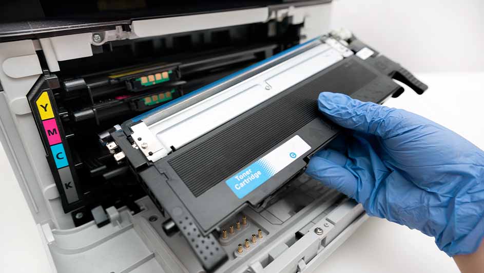 Een hand met een blauwe handschoen vervangt een zwarte tonercartridge in een printer met gele, roze en blauwe aanduidingen.