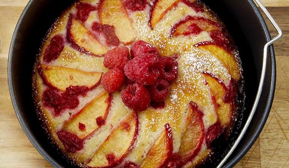 In a black baking tin lies a homemade tart with peach and raspberry, finished with icing sugar and fresh raspberries. Brand is not visible.