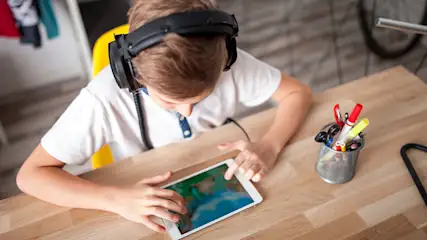 Een jongen met een koptelefoon gebruikt een tablet aan een houten bureau met pennen in een beker.