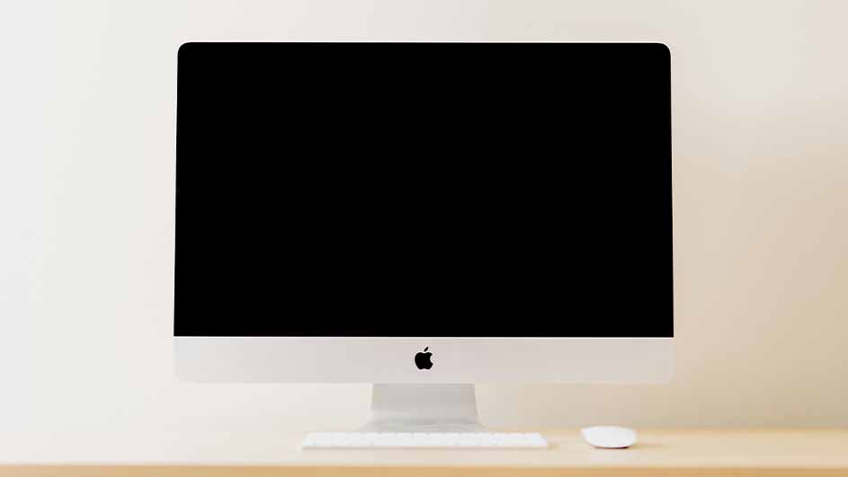 Een witte iMac met een zwart scherm staat op een houten bureau, met een wit toetsenbord en een witte muis ervoor.