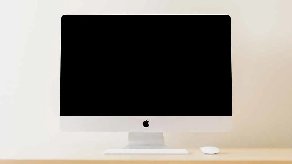 Een witte iMac met een zwart scherm staat op een houten bureau, met een wit toetsenbord en een witte muis ervoor.