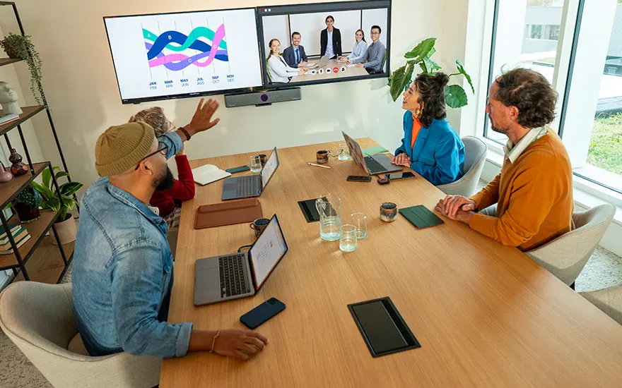 Vergaderruimte met collega's rond tafel, grote scherm toont videovergadering en kleurrijke grafieken voor hybride zakelijke presentatie.